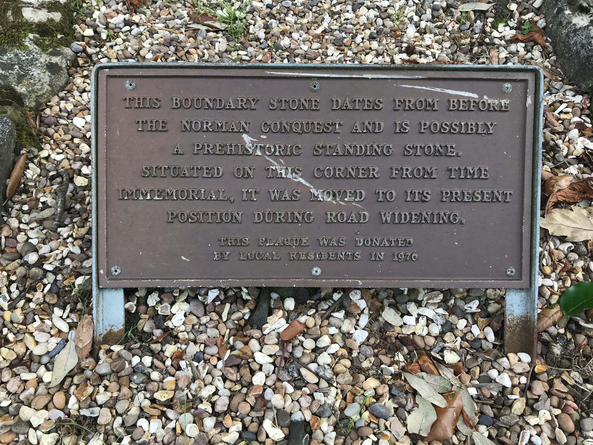 Rectangular plaque of brown-coloured bronze with embossed upper case writing, supported by two thin metal legs. Surrounded by gravel.